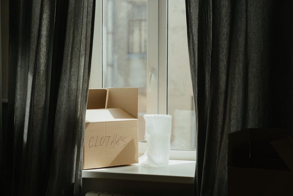 Inside a room with dark curtains on either side of a window, a cardboard box labeled 'CLOTHES' in black marker is positioned on the windowsill along with a neatly folded white fabric item, possibly a blanket or towel. The window is partially open, allowing natural light to illuminate the box and fabric, and revealing a blurred view of adjacent buildings outside. To the right of the window, a black piece of furniture, such as a chair or armrest, is partially visible. The setting suggests a home environment in the process of packing or preparing for a house move, with the cardboard box indicating ongoing packing activities by West Hampstead Removals as part of a home relocation or furniture transport service.
