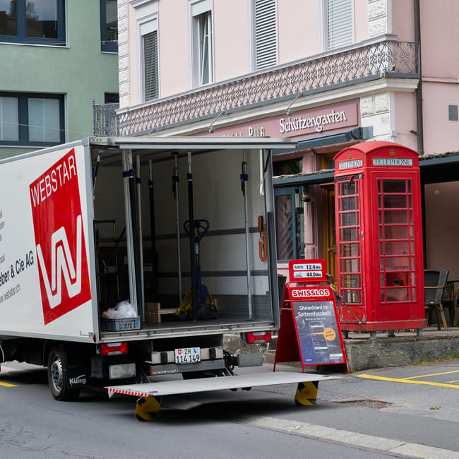 A white moving truck with an open rear door is parked on a city street in front of a building with a pink facade and balconies. Inside the truck, visible objects include two blue hand trucks, a yellow strap, and some packing materials. The truck's loading ramp is extended onto the pavement, and a pair of yellow and black wheel stoppers prevent movement. Nearby, there is a red British telephone booth positioned on the sidewalk close to a restaurant with a sign that reads 'Schutzzgarden.' The area appears prepared for a home relocation, with space cleared for furniture and boxes being loaded or unloaded, and the environment is well-lit by daylight. The scene suggests a local residential move, with furniture transport and packing in progress facilitated by West Hampstead Removals.