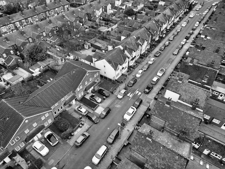 A black-and-white aerial photograph of a residential street with multiple houses and terraced properties, with parked cars lining both sides of the road. The street shows a mix of detached and semi-detached homes, some with front gardens and driveways, while others have on-street parking. In the foreground, a house with a tiled roof and a small front yard is visible, adjacent to the road where several vehicles are parked. Slightly further along the street, a van belonging to West Hampstead Removals is seen at the curbside, either in the process of loading or unloading household furniture and boxes wrapped in brown cardboard and plastic. The surrounding area consists of tightly packed rows of houses with tiled roofs, small backyards, and side alleys. Equipment such as trolleys or straps used for furniture transport might be visible nearby, and the overall scene captures the typical setting for house clearance and home relocation services, with minimal activity indicating an organized moving or packing process consistent with professional removals operations in Camden's Mill Lane NW6.
