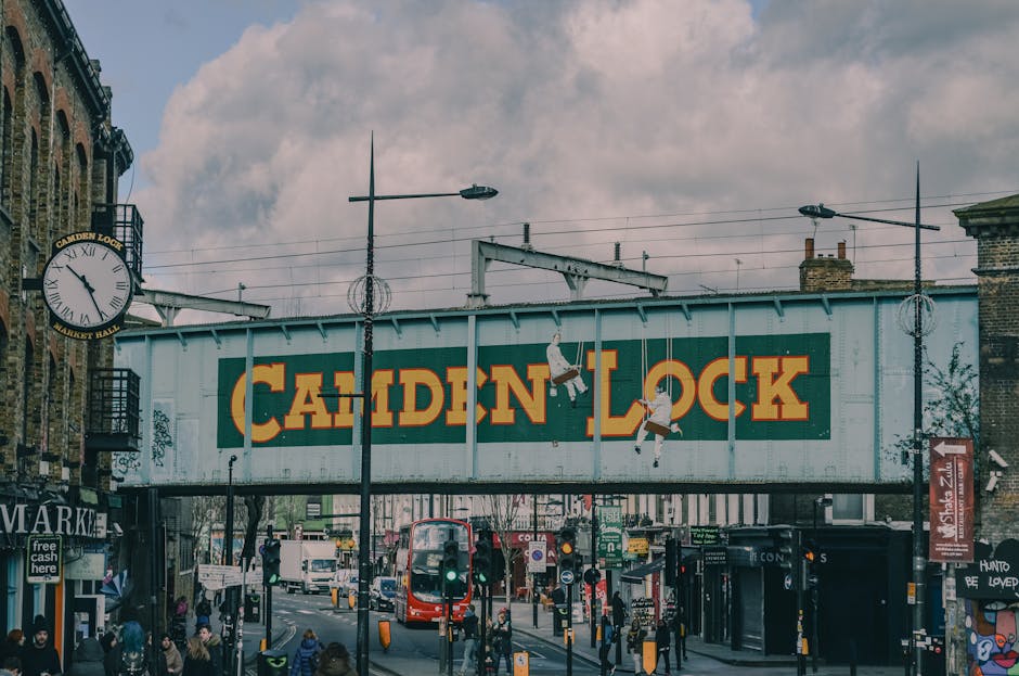 The image shows a street scene at Camden Lock with a large green and yellow sign hanging overhead, displaying 'CAMDEN LOCK' in bold yellow letters. The sign is mounted on a metal structure above the road, which also supports streetlights and electrical wiring. Below, there are various pedestrians walking along the pavement, some holding shopping bags, and a red double-decker bus moving through the street. Adjacent to the scene, buildings feature shopfronts, including a market and other local businesses. The sky is partly cloudy, casting natural light across the area. In the foreground, West Hampstead Removals is involved in a home relocation process, recently loading furniture and boxes onto their van outside a property on Mill Lane NW6. The furniture includes cardboard boxes, plastic-wrapped items, and a wooden chair, staged near the entrance for a nearby house clearance. Equipment such as trolleys and moving straps are visible as part of the loading process, which is executed with care and attention to safety, consistent with professional removals services supporting house clearance and furniture transport for residential moves in the Camden area.