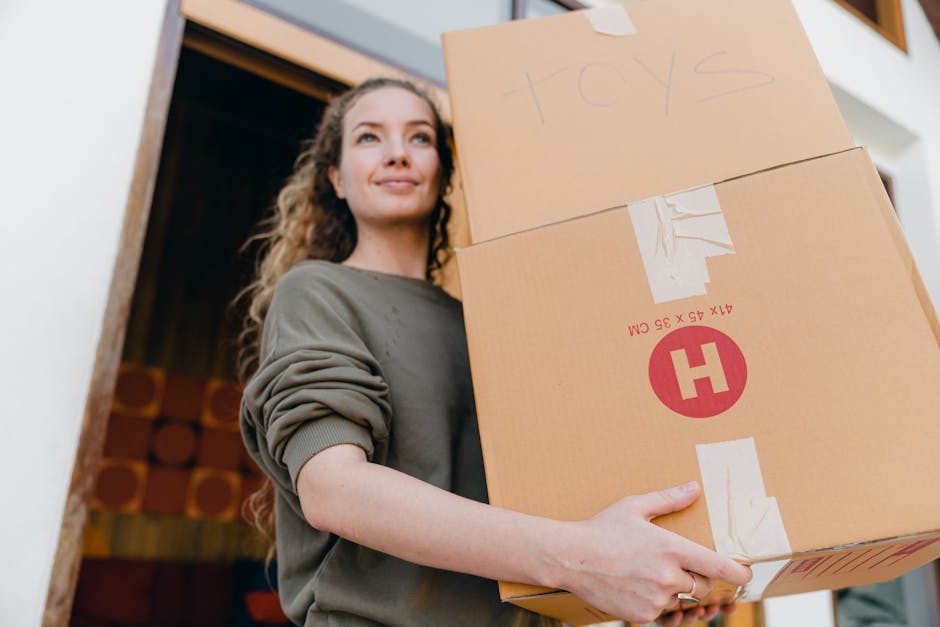 A professional mover from West Hampstead Removals, wearing a navy blue t-shirt with the company's logo, stands indoors next to a wall with a textured grey finish. He is holding a clipboard and using a