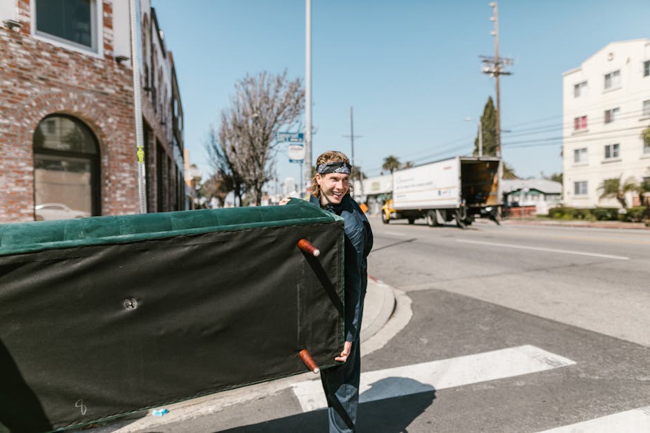 A person with a cheerful expression, wearing casual clothing and a headband, is walking outside on a city street during daytime while carrying a large, padded furniture transport bag with metal handles, used for home relocation. The bag is green with black sides and is positioned over their shoulder, indicating they are in the process of loading or unloading furniture for a move. In the background, a moving truck with an open rear door is parked on the side of the road, and residential buildings along with a few parked cars are visible. The environment includes clear weather with blue skies, and some trees are visible, one of which has no leaves, suggesting a transitional season. The scene reflects a furniture transport and packing process as part of a professional removals service, such as West Hampstead Removals, supporting efficient home relocation and moving logistics in NW6 Fortune Green area.