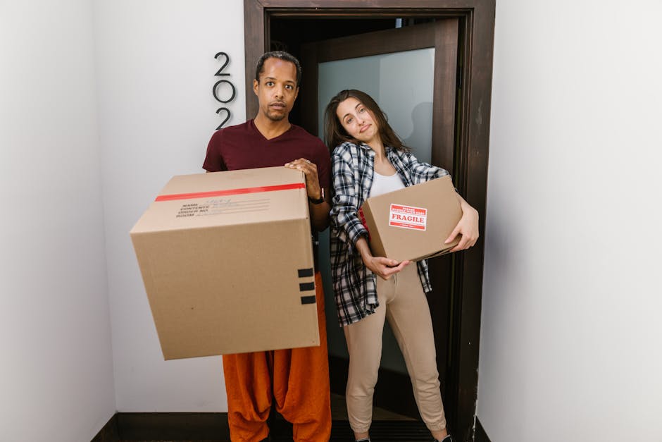 A man and a woman standing in a narrow internal hallway near a door, holding cardboard boxes during a home relocation process. The man, dressed in a maroon t-shirt and orange trousers, holds a large cardboard box in front of him, which has black packing tape and a shipping label. The woman, wearing a plaid shirt over a white top and beige trousers, leans slightly against the door frame while holding a smaller cardboard box marked with a red 'FRAGILE' label. The background features a doorway with a wooden door frame, and the walls are plain white, with the images capturing the loading or packing stage of a furniture transport and packing process for a house move. This scene aligns with house removals, emphasizing packing, loading, and object handling, as part of a professional moving service provided by West Hampstead Removals, supporting a smooth home relocation in NW6 Fortune Green.