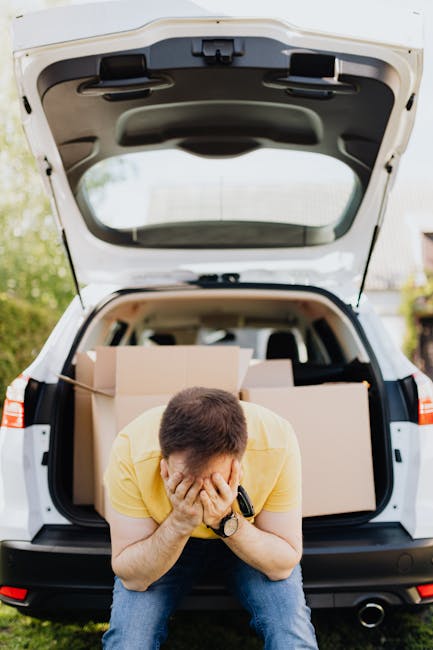 A man wearing a yellow T-shirt and blue jeans is crouched in front of an open black and white hatchback vehicle parked outdoors. He is holding his face with both hands, indicating frustration or fatig