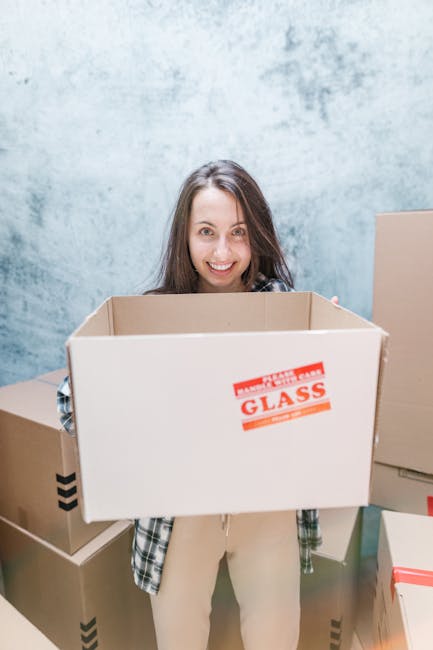A young woman with long brown hair smiling at the camera while holding an empty cardboard box labeled 'Glass' in red, inside a room with a light blue, slightly textured wall. She is wearing a checkere