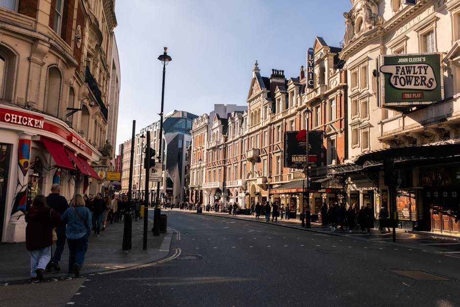 A busy street scene in a central urban area with a row of multi-story shops and commercial buildings on either side, featuring ornate architectural details and signage for venues such as 'Fawty Towers' and 'Lyric,' with pedestrians walking along the sidewalk, some engaged in shopping or conversations. In the foreground, a small group of people is walking past a storefront with a red awning labeled 'Chicken,' while others are waiting to cross at the pedestrian crossing. The street is lined with black lampposts and street signs, with a modern glass building visible further down the road. The scene is taken during daylight with clear skies, and the environment reflects a lively shopping district, typical of a London neighbourhood. West Hampstead Removals uses similar street scenes to illustrate urban house relocation and furniture transport activities, emphasizing the logistics involved in home moving services.