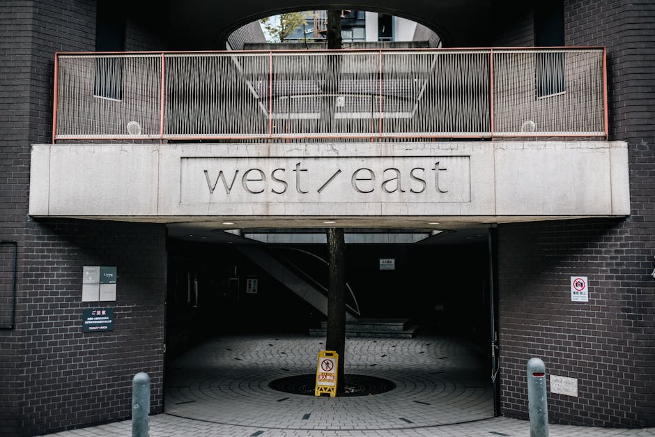 The image shows the exterior of a modern building with dark brick walls and an overhanging concrete balcony featuring a wire mesh railing in a peach or light rust colour. Underneath the balcony, there is an open driveway or passage used for home relocation activities, with a tree planted centrally on the paved pavement. Visible inside the driveway are material handling elements such as a wooden crate or packing box, along with a moving trolley with wheels used for furniture transport, and plastic wrapping materials possibly for furniture protection. There are a few objects positioned near the building’s entrance, including a metal bollard and a small yellow caution sign on the pavement, indicating ongoing loading or unloading activities. The area appears well-lit with natural daylight, suitable for moving services provided by West Hampstead Removals, as indicated by the signage on the building and the visible context of load carrying and furniture loading processes for house removals around West End Lane.