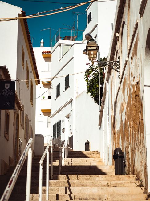 A narrow outdoor staircase constructed of stone with wooden steps, situated between white buildings featuring a mix of plaster and exposed brick walls on either side. A black trash bin is positioned at the bottom right of the stairs, while a traditional street lantern extends from a metal bracket attached to the right-side building. The sky is bright blue with no clouds, and various television antennas and rooftop structures are visible above the buildings. This scene illustrates the tight, winding staircases often found in NW6 buildings, which can present challenges during home relocation and furniture transport. West Hampstead Removals specializes in handling such narrow stair removals, ensuring careful packing, loading, and transport of household items in these confined spaces, supported by equipment like hand trucks and moving blankets to facilitate efficient moving processes within constrained urban environments.