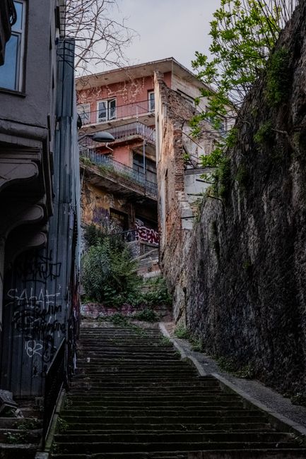 A narrow outdoor staircase made of concrete steps ascending between a graffiti-covered dark metal building on the left and a tall, weathered stone wall on the right, with sparse greenery and trees visible at the top. The staircase is located in a steep, urban setting typical of NW6 area buildings, and the scene is captured in natural daylight. This environment illustrates the challenges of navigating narrow stairways during home relocation or furniture transport, which West Hampstead Removals specializes in managing during house removals in the area.