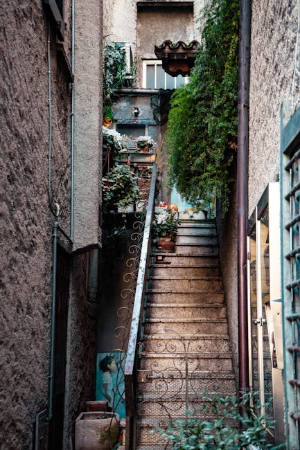 A narrow outdoor staircase constructed of stone with wooden steps, situated between white buildings featuring a mix of plaster and exposed brick walls on either side. A black trash bin is positioned at the bottom right of the stairs, while a traditional street lantern extends from a metal bracket attached to the right-side building. The sky is bright blue with no clouds, and various television antennas and rooftop structures are visible above the buildings. This scene illustrates the tight, winding staircases often found in NW6 buildings, which can present challenges during home relocation and furniture transport. West Hampstead Removals specializes in handling such narrow stair removals, ensuring careful packing, loading, and transport of household items in these confined spaces, supported by equipment like hand trucks and moving blankets to facilitate efficient moving processes within constrained urban environments.